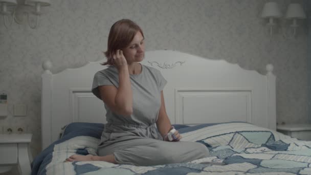 Mouvement lent des jeunes jolies femmes en robe grise mettant ses écouteurs sans fil et allumant la musique sur smartphone. Femme assise sur le lit avec téléphone dans les mains . — Video