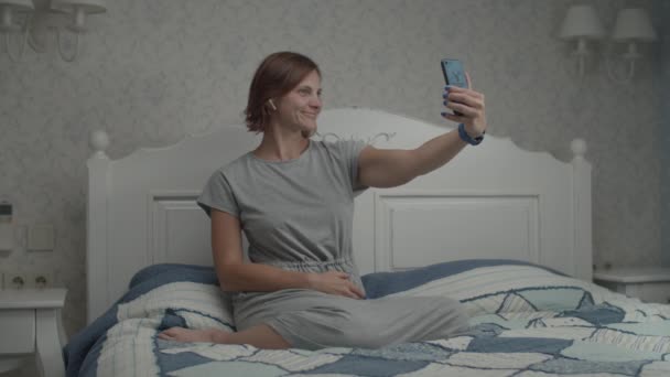 Zeitlupe junger hübscher Frauen in grauem Kleid und kabellosen Kopfhörern bewundert ihr Spiegelbild in der Smartphone-Kamera. Frau macht Selfie mit Handy im Schlafzimmer — Stockvideo