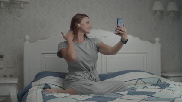 Αργή κίνηση των νεαρών όμορφων γυναικών με γκρι φόρεμα και ασύρματα ακουστικά θαυμάζει την αντανάκλασή της στην κάμερα smartphone. Γυναίκα λήψη selfie στο τηλέφωνο στο υπνοδωμάτιο — Αρχείο Βίντεο