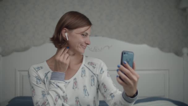 Pijamalı ve kablosuz kulaklıklı kadınlar akıllı telefon kamerasındaki yansımasına hayran. Kadın yatak odasında telefonla selfie çekiyor. — Stok video