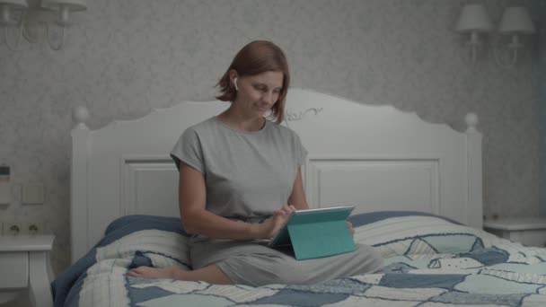 Slow motion van jonge mooie vrouwen in grijze jurk surfen op internet met haar tablet computer en draadloze oortelefoons zitten op het bed. — Stockvideo