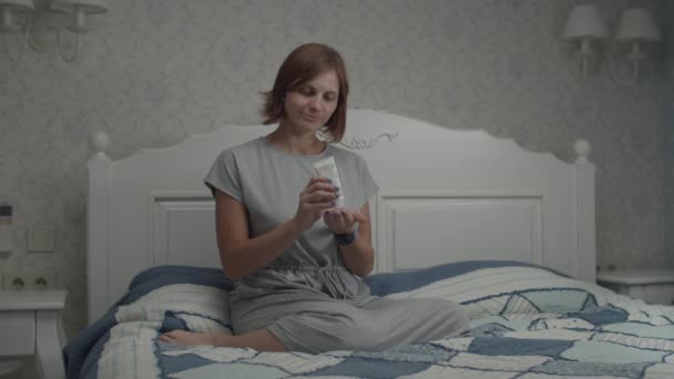 Langzame beweging van jonge mooie vrouwen in grijze jurk aanbrengen van vocht crème op haar gezicht zittend op het bed in Provençaalse interieur. — Stockvideo