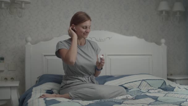 Mouvement lent des jeunes jolies femmes en robe grise mettant ses écouteurs sans fil et allumant la musique sur smartphone. Femme assise sur le lit avec téléphone dans les mains . — Video