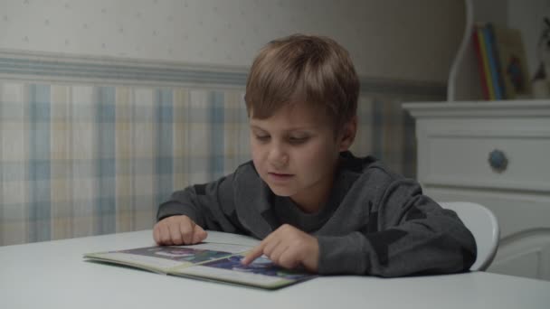 Rapaz autista a ler livros e sentado à mesa em câmara lenta. Um miúdo com autismo a olhar para o livro. Consciência do autismo — Vídeo de Stock