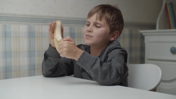 Niño autista pelando un plátano en cámara lenta sentado en la mesa. El chico está contento con la comida. Conciencia del autismo — Vídeo de stock