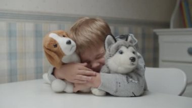 Sarışın çocuk, yumuşak yavru köpeğe sarılıyor ve ağır çekimde masada oturuyor. Çocuk, yumuşak köpek oyuncaklarıyla oynamaktan mutlu.. 
