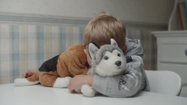 Niño rubio abrazando juguete suave cachorro sentado en la mesa en cámara lenta. Niño está feliz de jugar con juguetes de perro suave . — Vídeo de stock