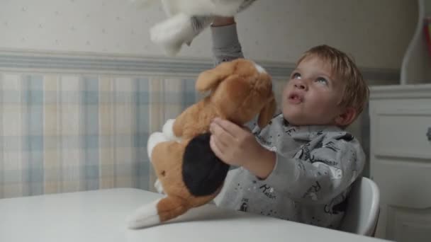 Szőke fiú játszik puha kiskutya játék ül az asztalnál lassított felvételen. Gyermek boldog játszani és ölelés puha kutya játékok. — Stock videók