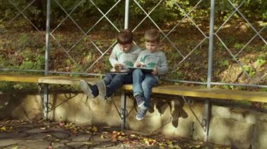 Sonbahar parkında elinde aletlerle bankta oturan iki kardeş. Sonbahar parkında akıllı telefon ve tablet bilgisayarla oynayan çocuklar. Yavaş çekimde sabit çekimde çek 