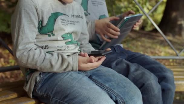 Dva bratři sedí na lavičce v podzimním parku s pomůckami. Kluci hrajou na smartphonu a tabletu v podzimním parku. Střílet na steadicam ve zpomaleném filmu — Stock video