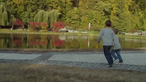 Dois irmãos correndo em belo parque de outono junto ao lago em câmera lenta. Rapazes a brincar e a correr no parque de outono. Atire com steadicam . — Vídeo de Stock