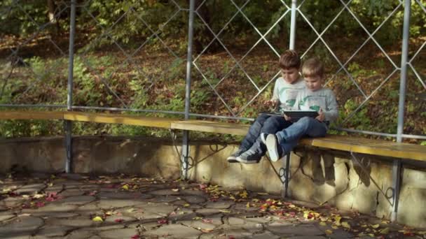 Twee broers zitten op de bank in het najaarspark met gadgets. Jongens spelen op smartphone en tablet computer in het najaar park. Schiet op standvastig in slow motion — Stockvideo