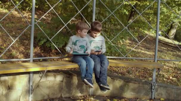 Due fratelli seduti sulla panchina nel parco autunnale con gadget. Ragazzi che giocano su smartphone e tablet in autunno parco. Spara su steadicam al rallentatore — Video Stock