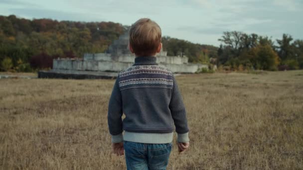 Bambino che cammina sul campo autunnale con struttura in cemento sullo sfondo. Spara al rallentatore, steadicam . — Video Stock