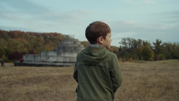 Kleine jongen loopt op het herfstveld met betonnen structuur op de achtergrond. Schiet in slow motion, Steadicam. — Stockvideo