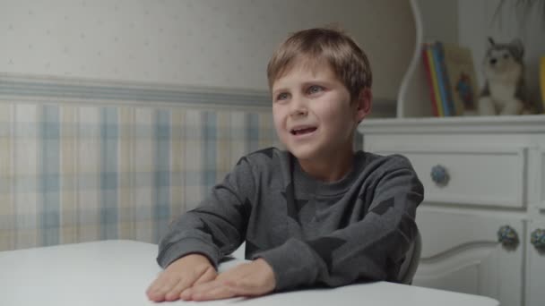 Аутистический мальчик жестикулирует руками, сидя за столом в замедленной съемке. Счастливый ребенок с аутизмом пытается говорить. Осознание аутизма — стоковое видео