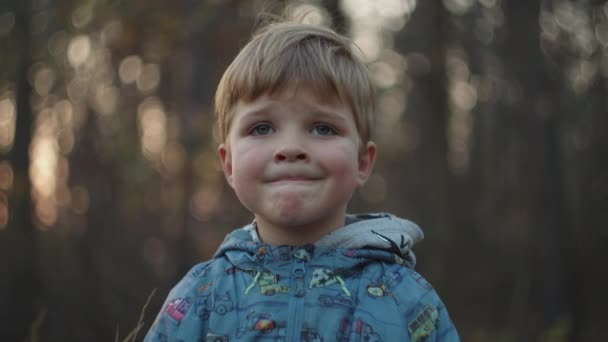 Günbatımı sonbahar parkında gülümsemeye çalışan üzgün bir çocuk. Sarışın çocuk sonbahar ormanında ağır çekimde gülümsüyor.. — Stok video