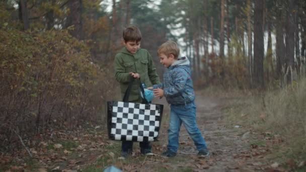 Two boys in jackets collecting plastic waste in fall park on sunset in slow motion. Kids are happy to find recyclable garbage in forest. — Stock Video