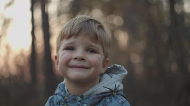 Günbatımı sonbahar parkında gülümsemeye çalışan üzgün bir çocuk. Sarışın çocuk sonbahar ormanında ağır çekimde gülümsüyor.. — Stok video