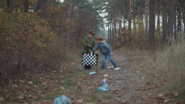 Deux garçons en vestes ramassant des déchets plastiques dans le parc d'automne au coucher du soleil au ralenti. Les enfants sont heureux de trouver des déchets recyclables dans la forêt . — Video