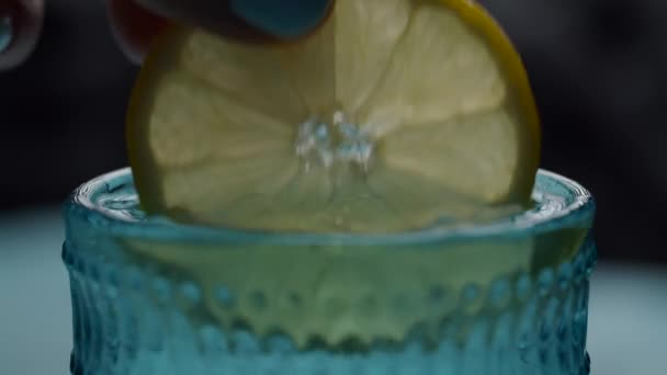 Zavřít citrónový plátek v ženské ruce namáčení ve vodě a kostky ledu v modrém skle. Kulatý citrónový kousek s nápojem ve zpomaleném makro pohledu. 