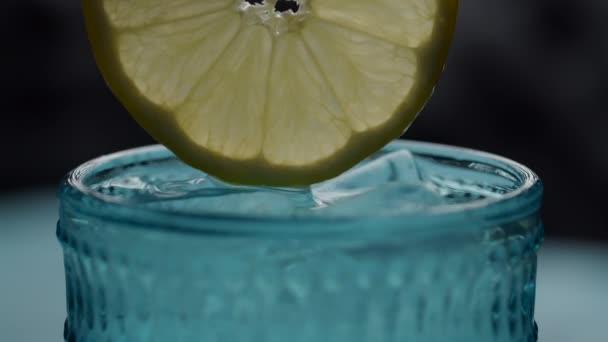 Chiudere di fetta di limone in mano femminile lasciando cadere in acqua e cubetti di ghiaccio in vetro blu. Pezzo rotondo di limone in bevanda al rallentatore macro vista . — Video Stock