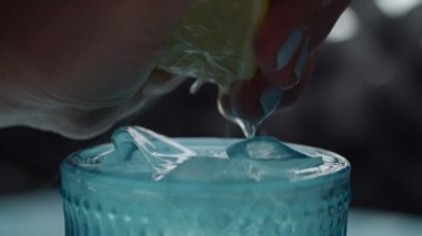 Kadın elinin buzlu mavi bir bardak içeceğe limon suyu sıkması gibi. Limon meyvelerinin yarısı yavaş çekim makro görüntüsünde sıkışır.