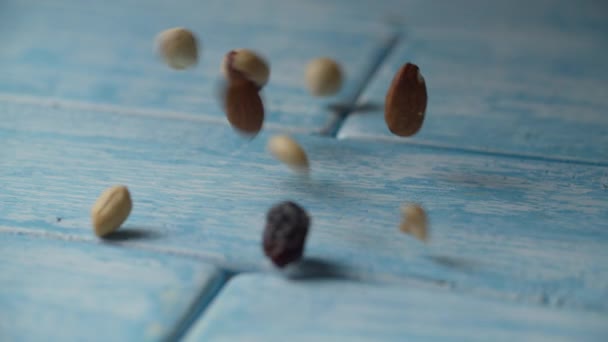 Nueces saladas mixtas y pasas que caen sobre la mesa azul en cámara lenta, vista macro. Almendras, avellanas, anacardos, cacahuetes y pasas que caen sobre la superficie brillante . — Vídeos de Stock