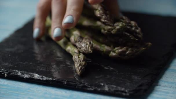 Mano de mujer poniendo manojo de espárragos verdes frescos en la placa negra, vista macro en cámara lenta. Primer plano de espárragos verdes vegetales en manos femeninas . — Vídeos de Stock