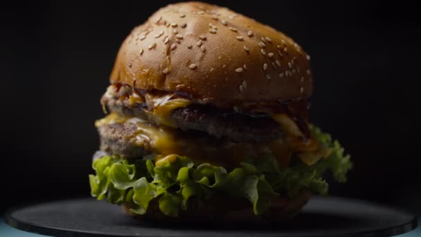 Finom burger dupla marhahússal, olvasztott sajttal, salátával és zöldséggel, sötét alapon forgatva, lassított felvételen. — Stock videók