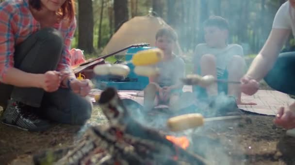 Dwie matki gotują kiełbaski i kukurydzę w ogniu, podczas gdy dwóch synów siedzi na kocu piknikowym podczas letnich wakacji rodzinnych z namiotem w lesie. Zwolnij, Steadicam shot. — Wideo stockowe