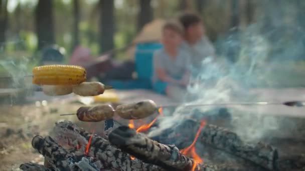 Zbliżenie kiełbasy i kukurydzy na szaszłyki są gotowane w ogniu z dymem podczas letnich rodzinnych wakacji kempingowych z namiotem w lesie. Zwolnij, Steadicam shot. — Wideo stockowe