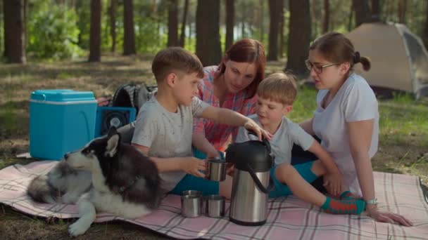 Two moms and two sons drinking tea on picnic blanket during summer family camping vacation with tent in forest. Boy pouring hot drink from thermos. Slow motion, steadicam shot. — Stock Video