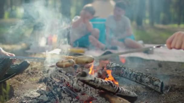 Zbliżenie kiełbasy i kukurydzy na szaszłyki są gotowane w ogniu z dymem podczas letnich rodzinnych wakacji kempingowych z namiotem w lesie. Zwolnij, Steadicam shot. — Wideo stockowe