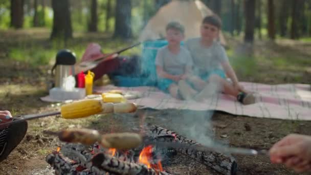 Близько сосисок і кукурудзи на шампурах готуються під час літнього сімейного кемпінгу з наметом у лісі. Повільний рух, стрімкий постріл . — стокове відео