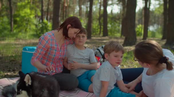 Dos madres, dos hijos y un perro Husky siberiano descansando sobre una manta de picnic durante las vacaciones de campamento familiar de verano con tienda en el bosque. cámara lenta, disparo steadicam . — Vídeo de stock