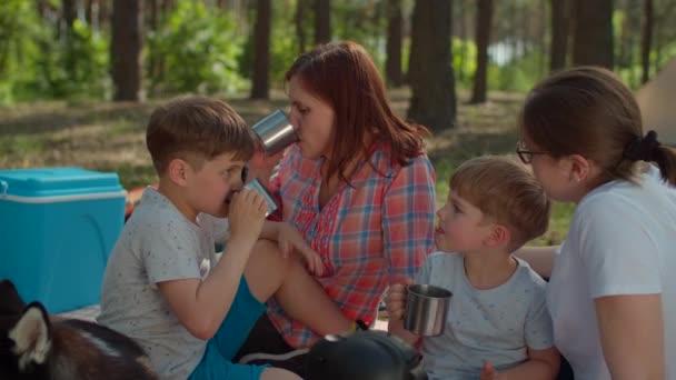 Twee moeders en twee zoons drinken thee op een picknickdeken tijdens de zomervakantie met tent in het bos. Gelukkige familie met hond. Langzame beweging, standvastig schot.. — Stockvideo