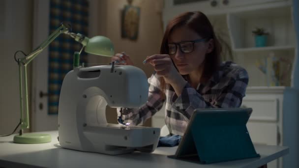 30s γυναίκα μαθαίνει σε απευθείας σύνδεση πώς να ράψει στη ραπτομηχανή από τον υπολογιστή tablet κάθεται στο σπίτι. Νεαρή μητέρα ράψιμο παιδικά ρούχα με βοήθεια στο διαδίκτυο. — Αρχείο Βίντεο