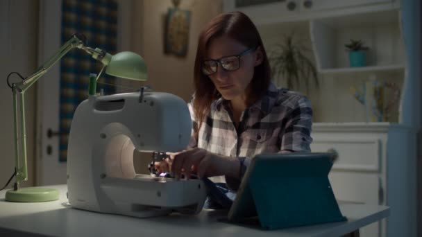 30 років жінка вчить онлайн шити машину за допомогою планшетного комп'ютера, сидячи вдома. Молода мама шиє дитячий одяг за допомогою Інтернету . — стокове відео