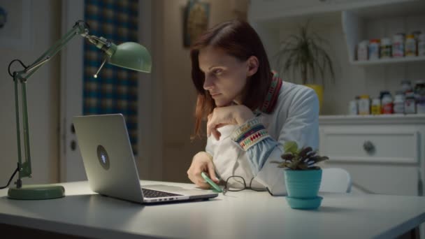 Jovem de 30 anos médica preparando-se para trabalhar em casa online. Mulher de casaco médico branco com estetoscópio no pescoço usando óculos e laptop de abertura para o trabalho . — Vídeo de Stock