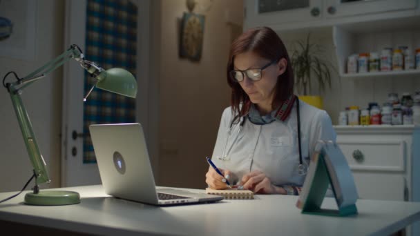 Pracuje w domu 30-tki, lekarz ma konsultacje online z pacjentem. Kobieta w białym płaszczu lekarza ze stetoskopem na szyi rozmowy wideo będzie chory pacjent. — Wideo stockowe