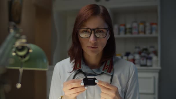 Travailler à domicile 30s femme médecin montre en ligne comment utiliser oxymètre de pouls. Femme en manteau de médecin blanc avec stéthoscope sur le cou parlant à la caméra. — Video