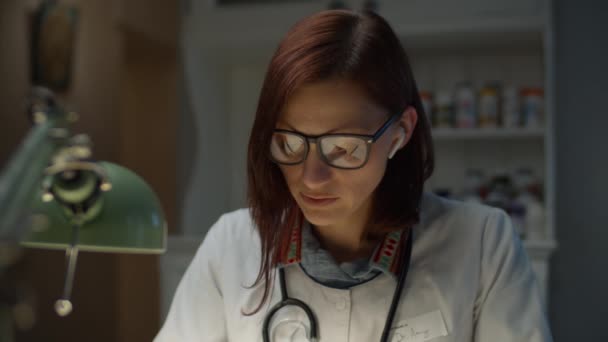 Young 30s female doctor making notes. Woman in white doctor coat with stethoscope on neck writing and talking looking at camera. — Stock Video