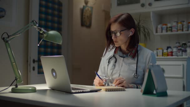 Молодая 30-летняя женщина-врач разговаривает онлайн с больным пациентом и делает заметки. Женщина в белом халате с стетоскопом на шее работает с помощью ноутбука и интернета . — стоковое видео