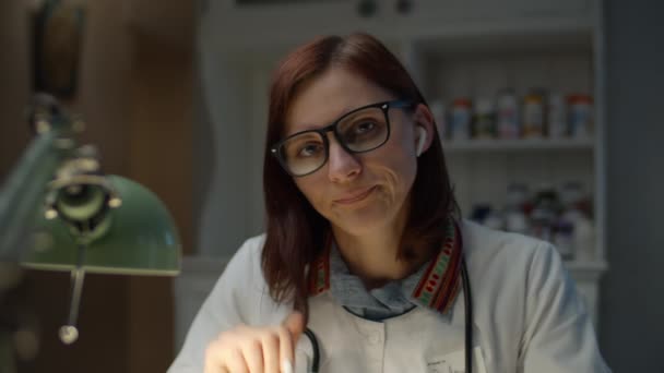 Jeune femme médecin trentenaire finit de parler en ligne. Femme fatiguée en manteau de médecin blanc avec stéthoscope sur le cou a mal de tête après la journée de travail. — Video