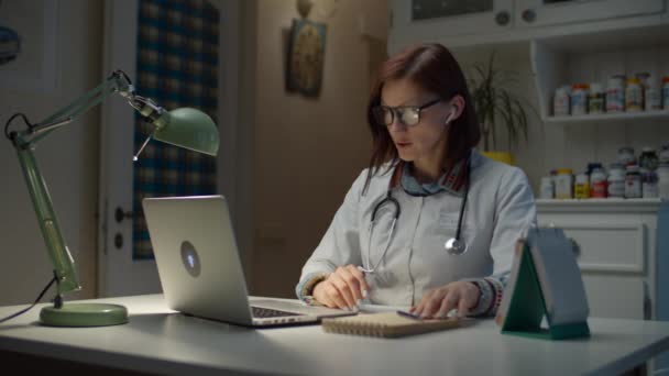 Unga 30-talet kvinnlig läkare talar online med sjuka patienten och göra anteckningar. Kvinna i vit läkare rock med stetoskop på halsen arbetar med bärbar dator och internet. — Stockvideo