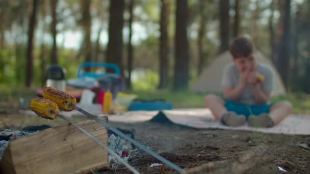 Portrait of school boy eating grilled corn holding in hands during summer family camping vacation with tent in forest. Focus on fire with cooking corn in front view. Slow motion, steadicam shot. — Stock Video