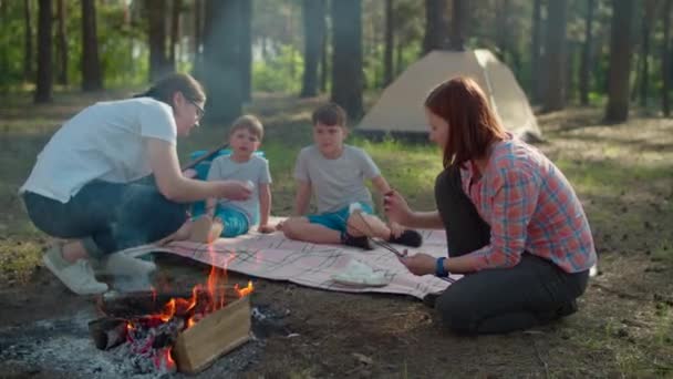 Lycklig familj med två mödrar och två söner som lagar marshmallows. Två kvinnor och två pojkar som har sommarcampingsemester i skogen. Slow motion, steadicam skott. — Stockvideo