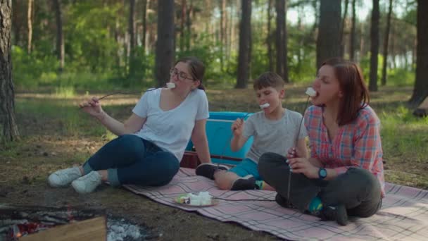 Dos madres y su hijo comiendo malvaviscos en palos. Dos mujeres y dos niños teniendo vacaciones de campamento de verano en el bosque. cámara lenta, disparo steadicam . — Vídeo de stock