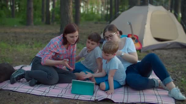 Two 30s mothers and two sons watching tablet computer laying on picnic blanket on summer camping vacation with tent in forest. Happy family with Siberian Husky dog. Slow motion, steadicam shot.